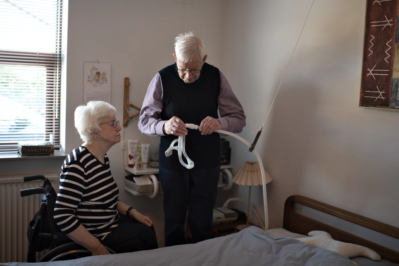 Kvinde sidder i koerestol og kigger paa mand der staar med CPAP-maske